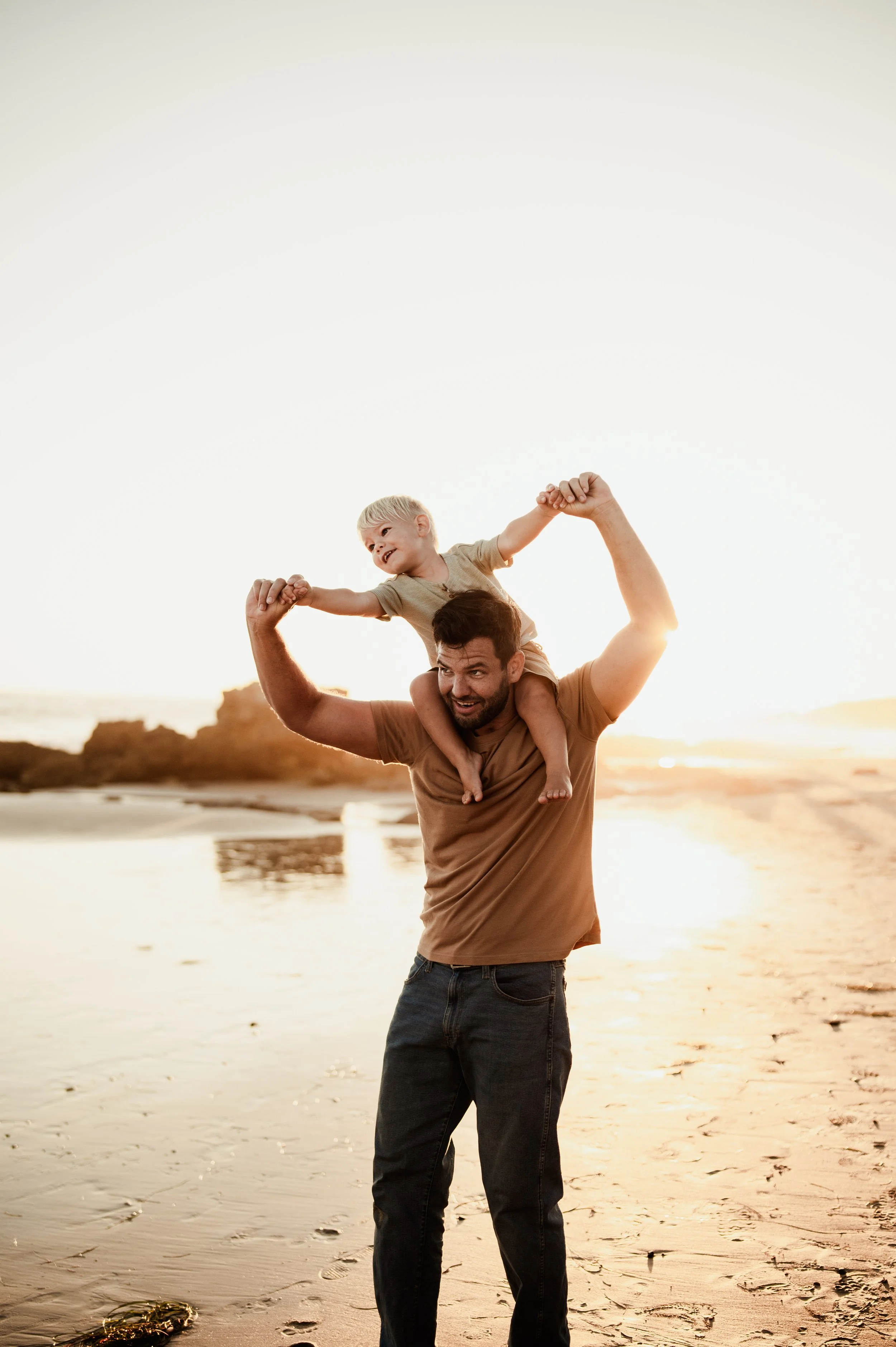 A man carrying a young boy on his shoulders on a beach at sunset, with rocky cliffs in the background.