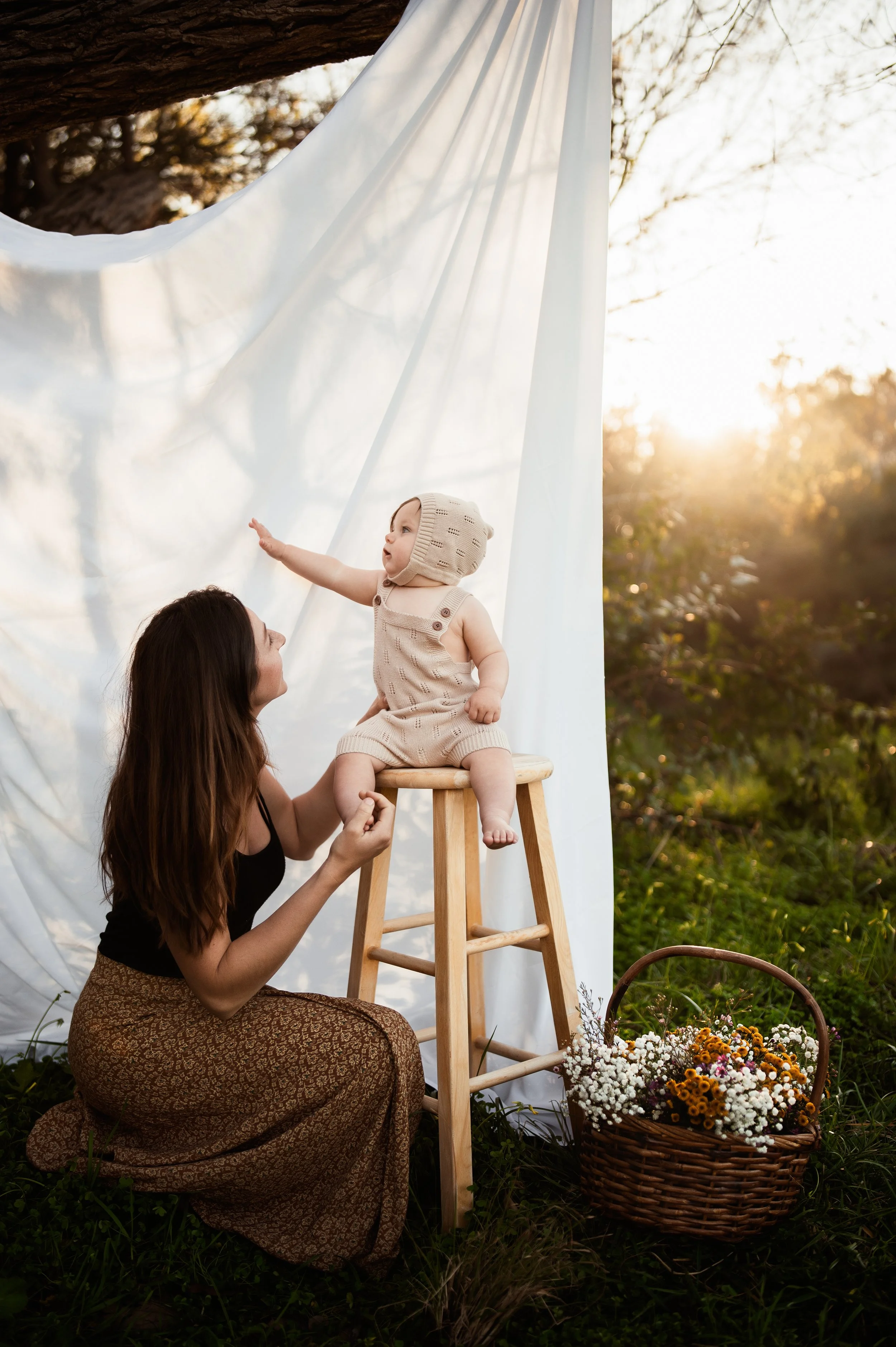 Woman kneeling beside a smiling baby sitting on a wooden stool outdoors with white fabric backdrop and sunlight. Basket of flowers on the grass nearby.