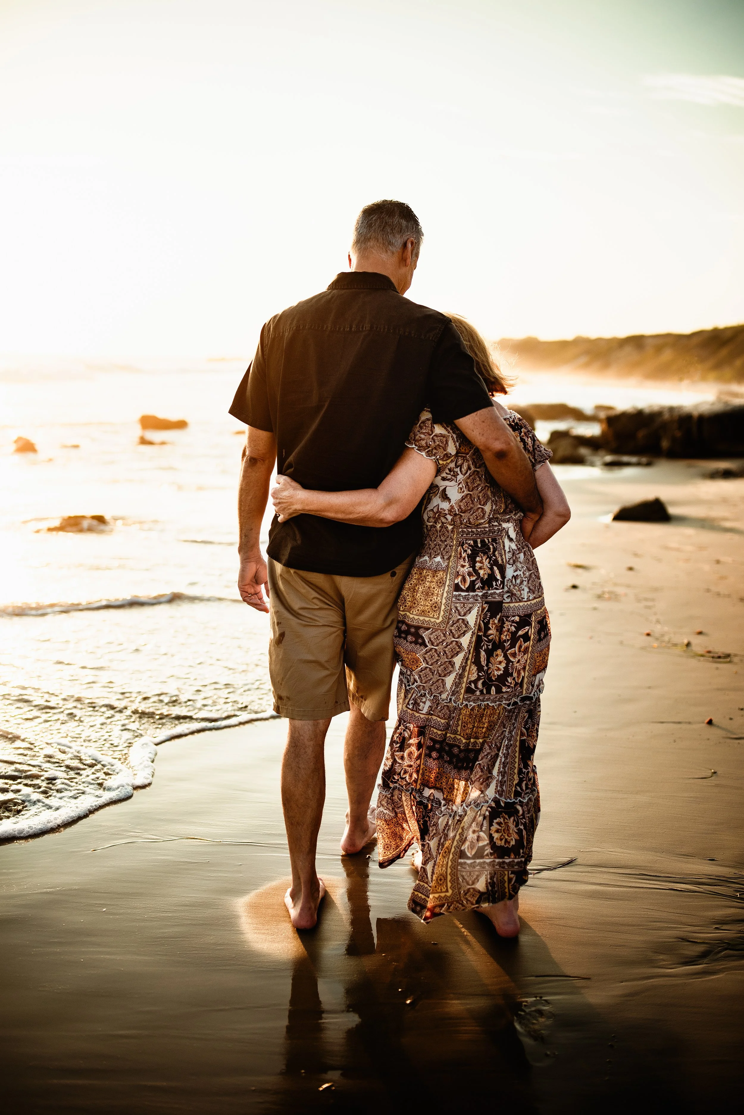 Couple walking on a beach at sunset, embracing, with waves and sand visible.