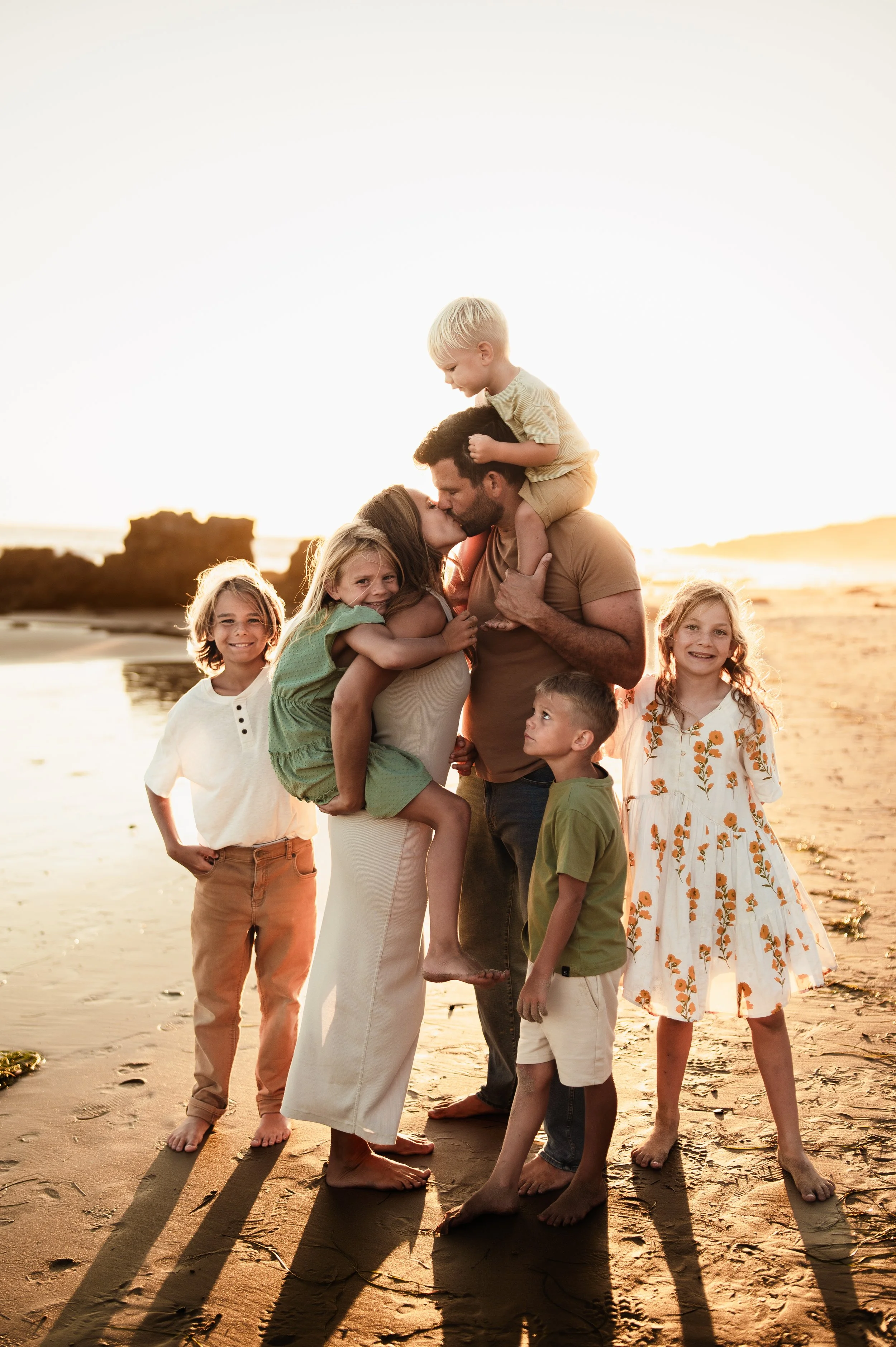 A family of seven at the beach during sunset, with some members hugging, kissing, and standing in a relaxed pose on the sand.