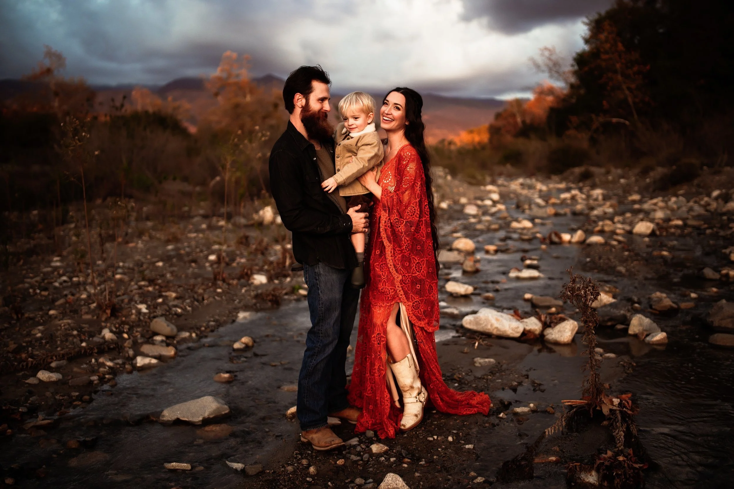 Family of three standing by a rocky stream, smiling, with mountains and cloudy sky in the background. A man in dark clothing holds a child, while a woman in a red dress stands beside them.