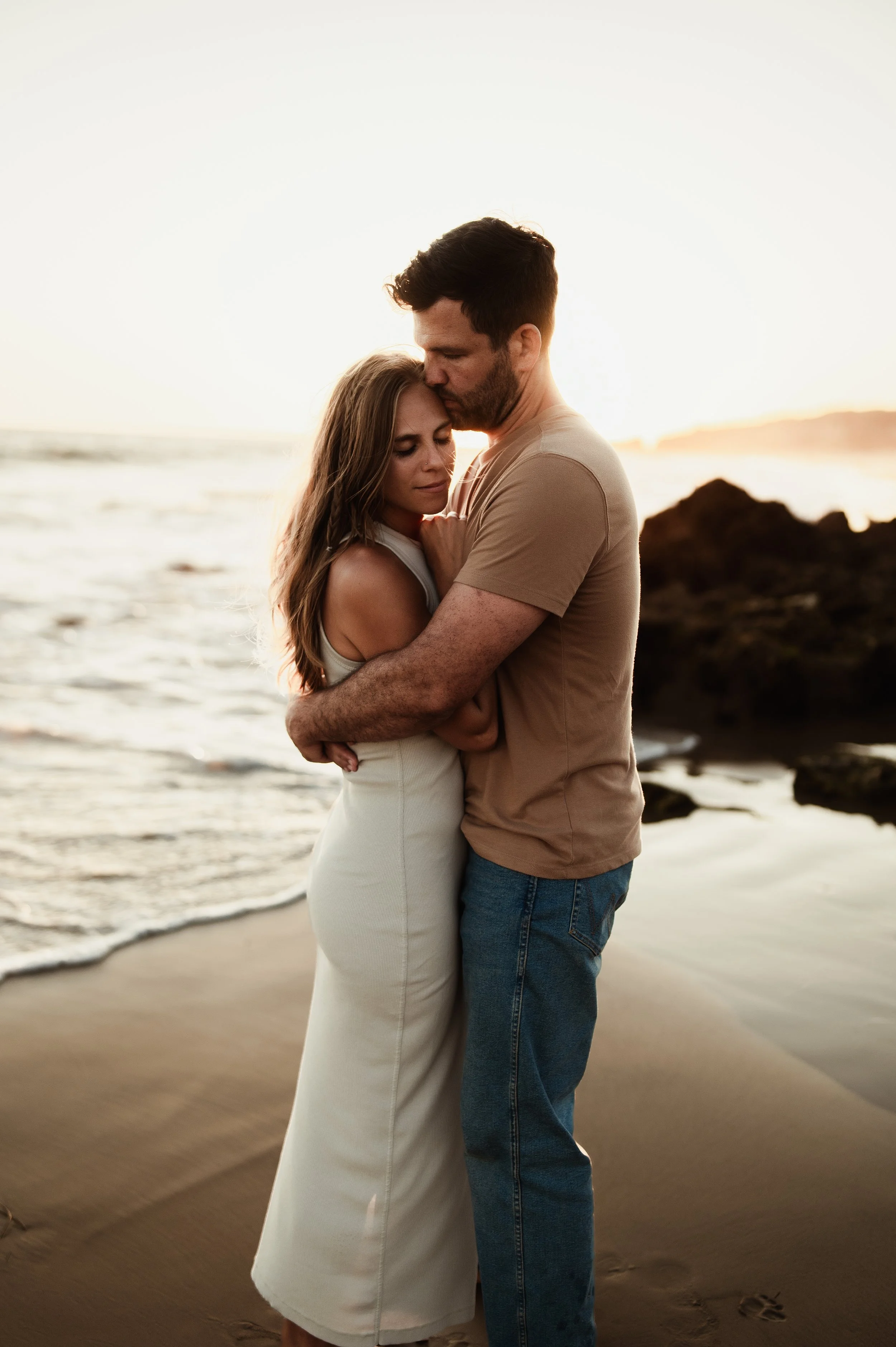 A man and woman embracing on a sandy beach during sunset, with waves and rocks in the background.