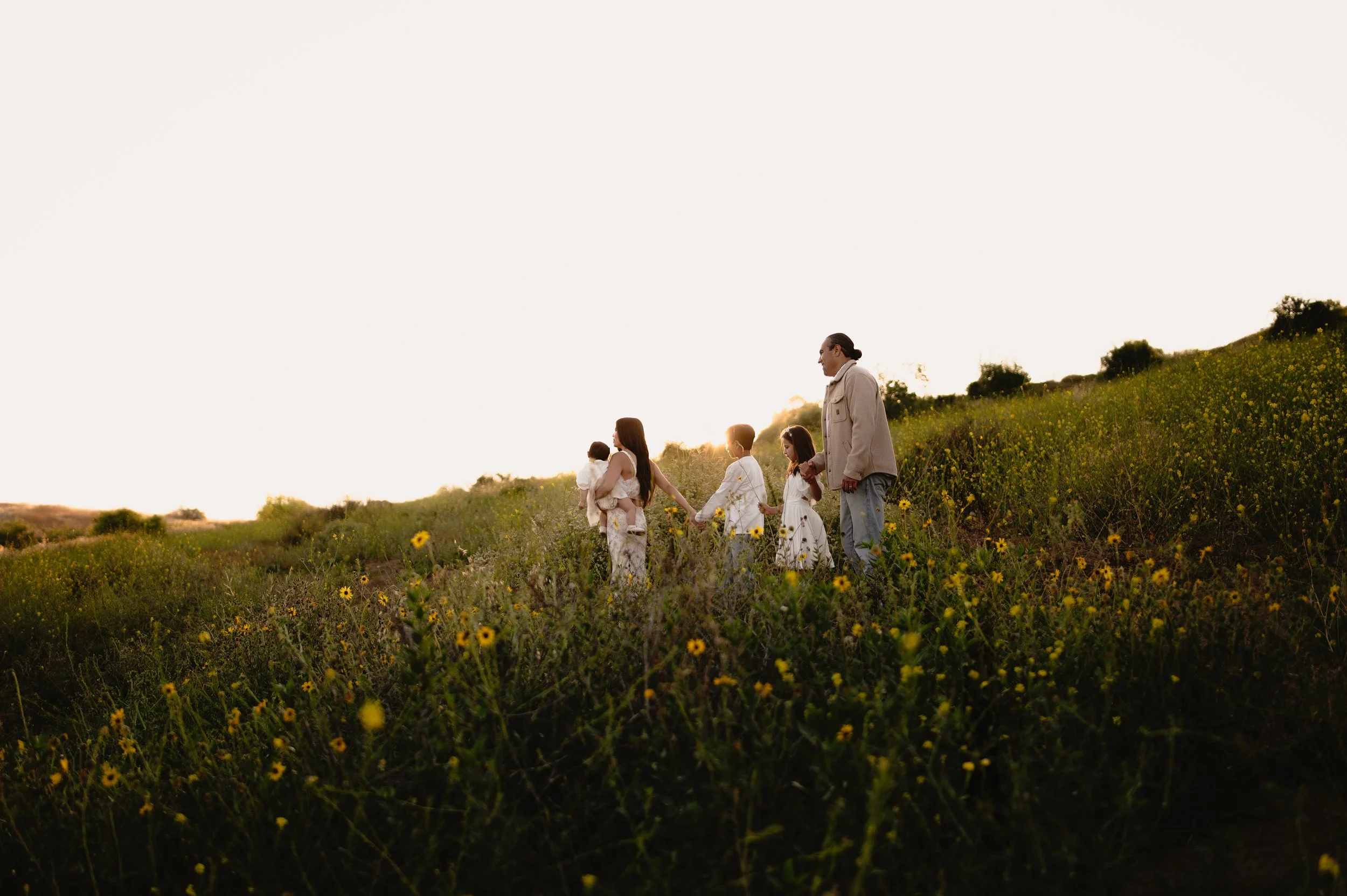 A family holding hands and walking through a field of yellow wildflowers at sunset, with a person wearing headphones.