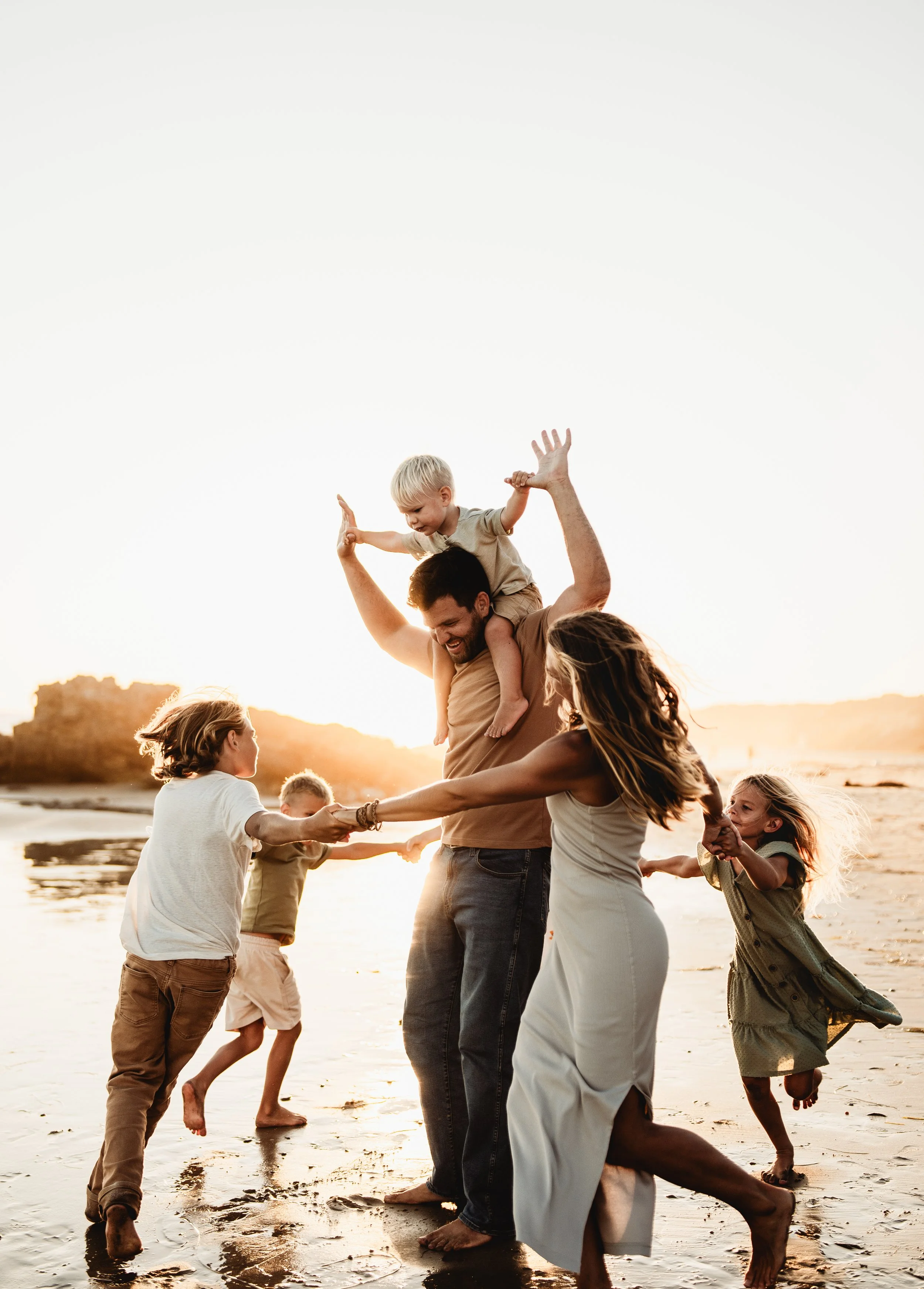 A group of children and adults playing and holding hands on the beach at sunset, with a man carrying a young boy on his shoulders, all smiling and enjoying the moment.
