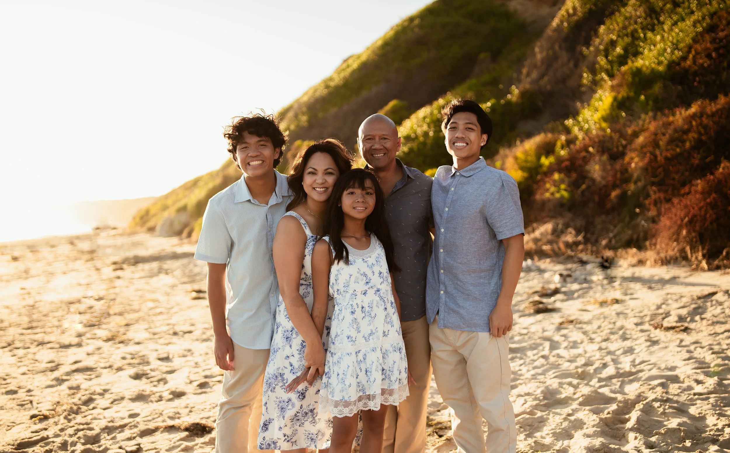 Family of five with teenagers, posing at crystal cove state beach in Newport. Standing on the sand with cliffs behind them at sunset.