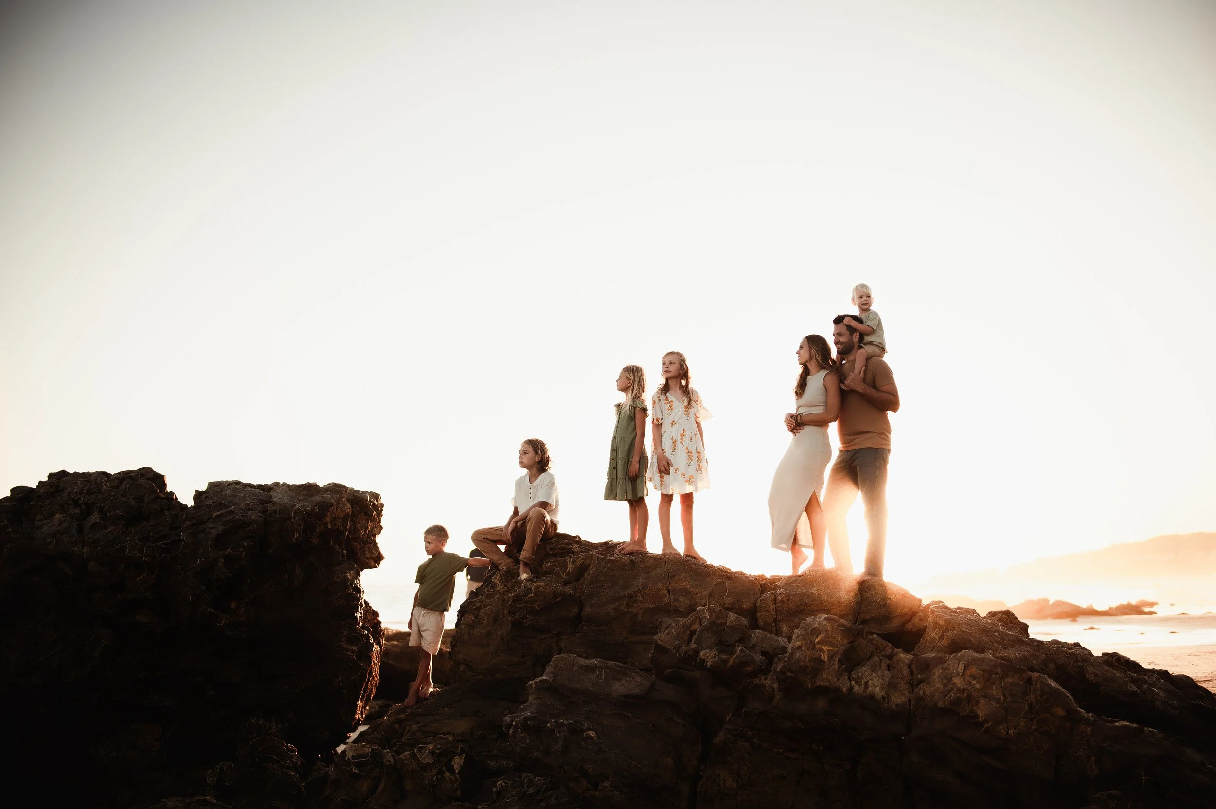 Family of seven standing and sitting on rocks by the water at sunset, with one man carrying a child on his shoulders, and the others looking at the horizon.