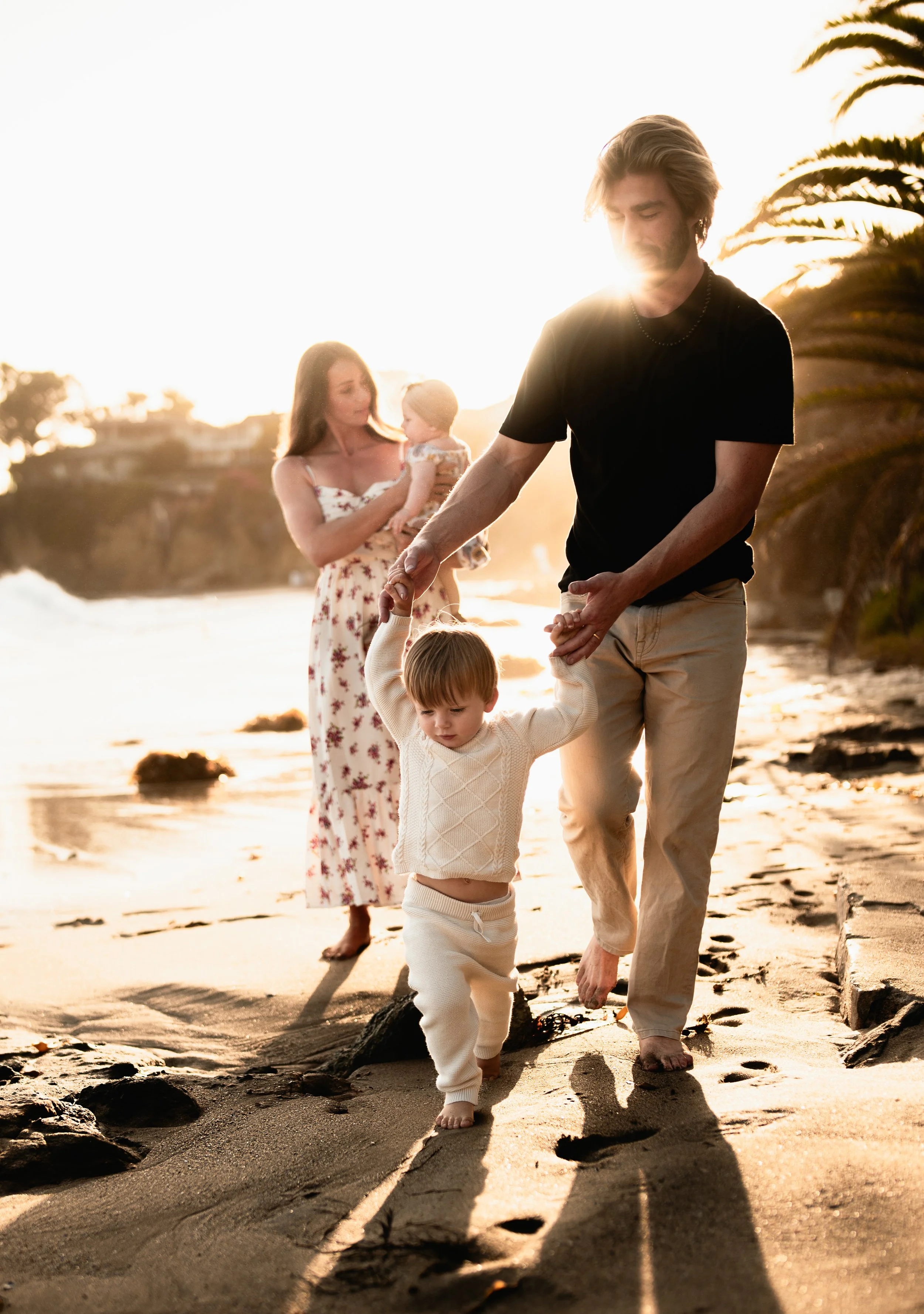 a father helps his son walk along the beach during sunset while the mom holds baby girl
