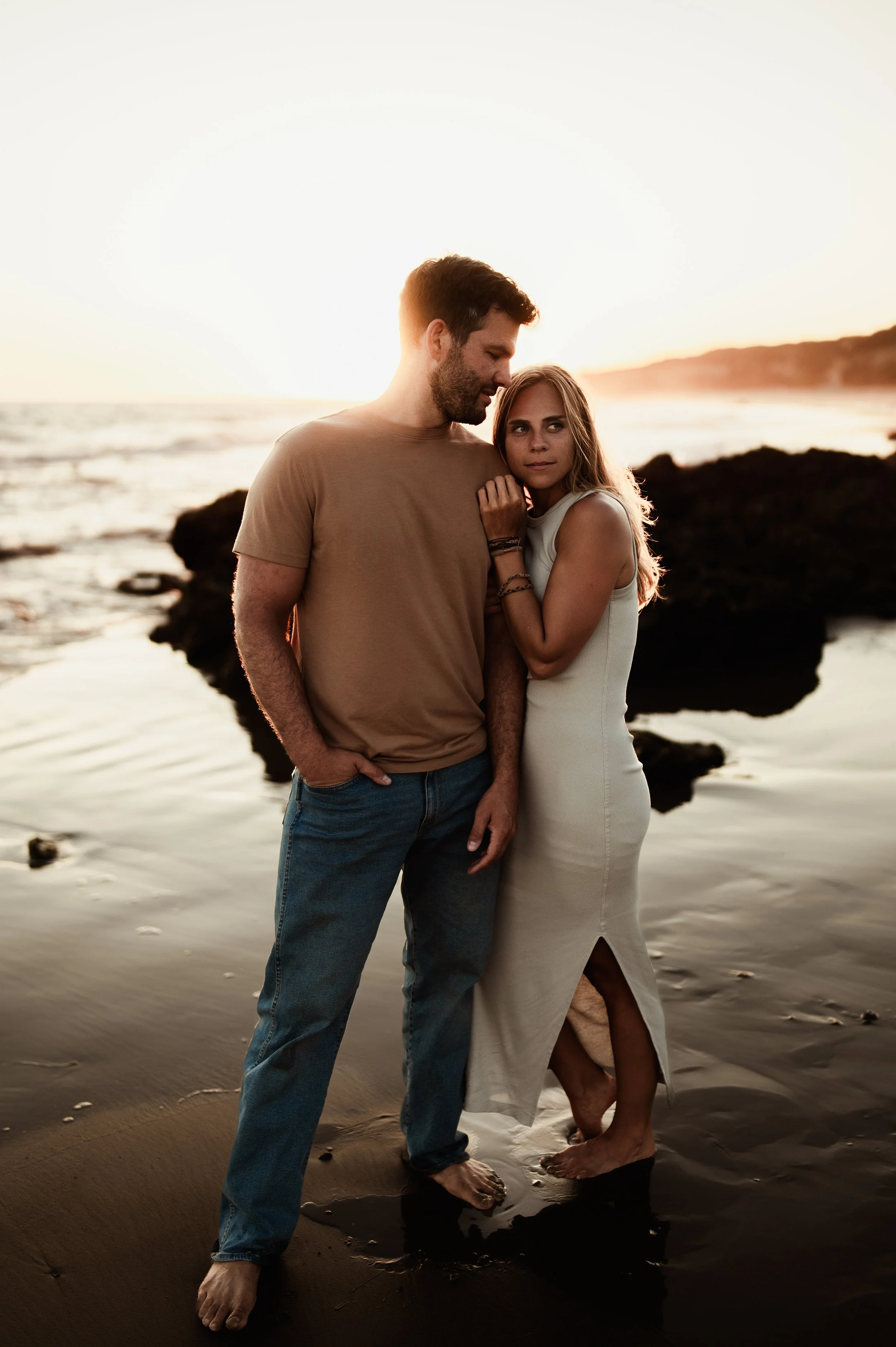 A man and woman standing close together on a beach at sunset, holding hands and barefoot, with ocean waves and rocks in the background.