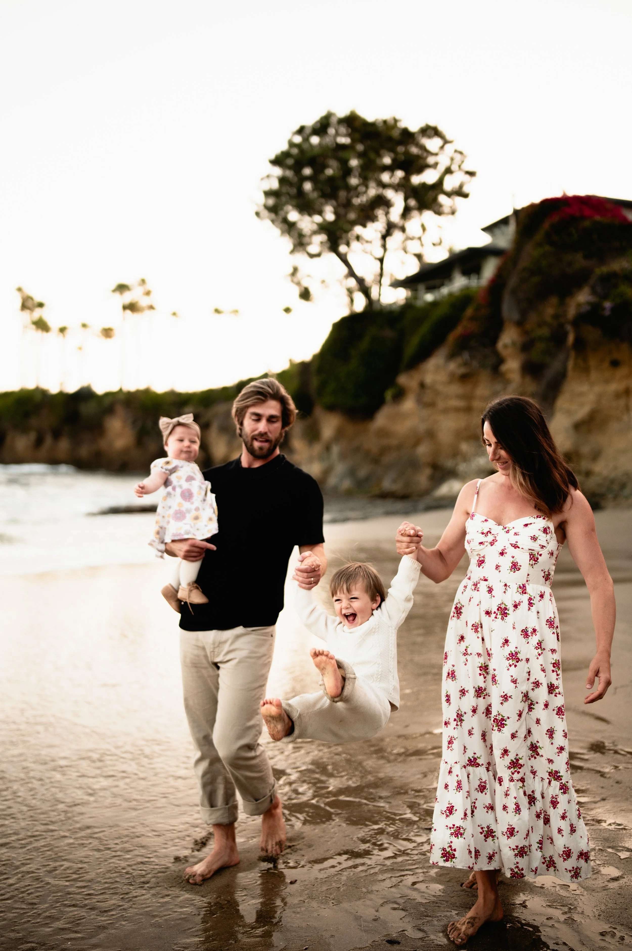 a mother and father play with their laughing son and daughter at the beach with cliffs in the background