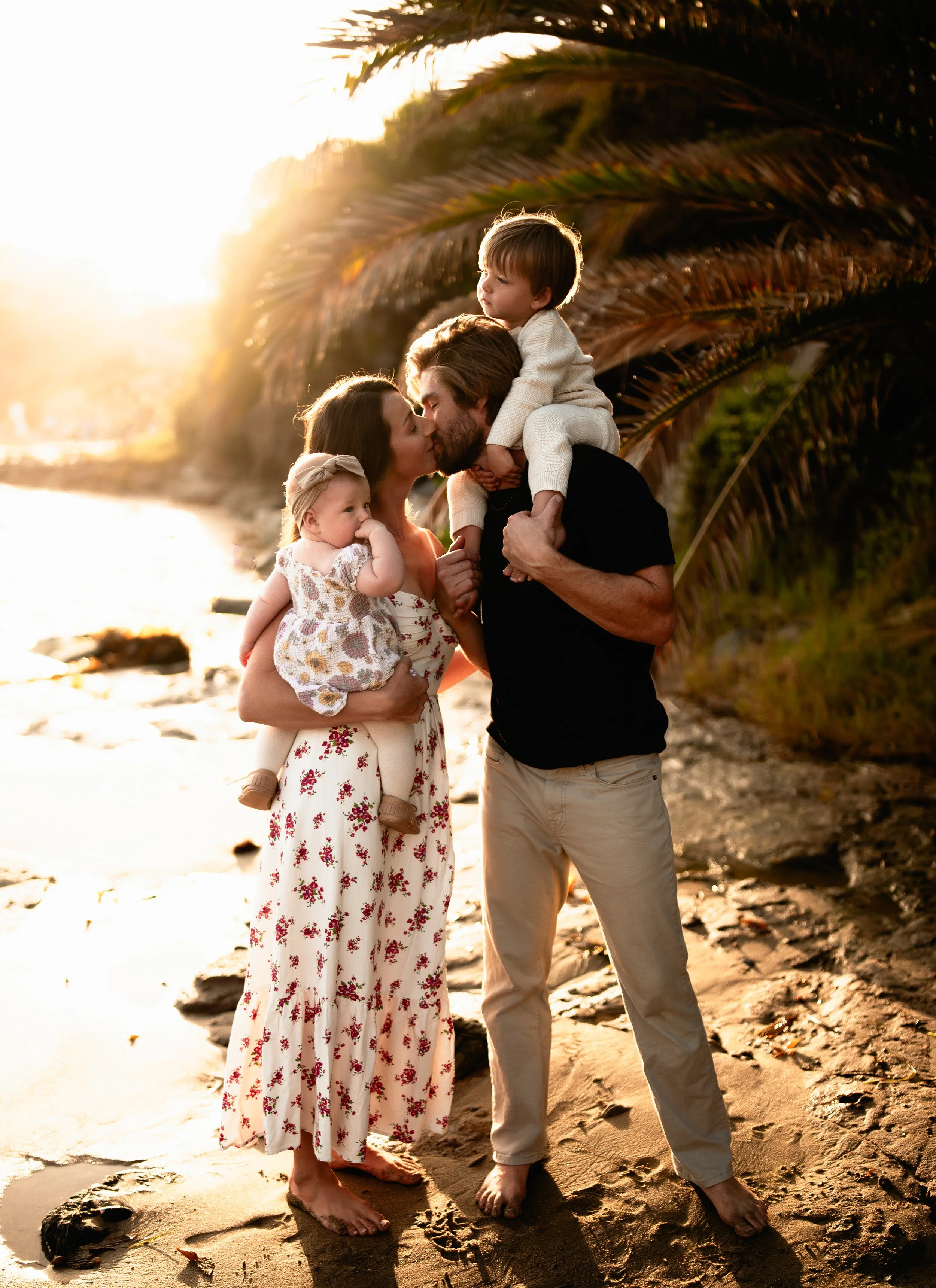 A mother and father kiss during sunset at the beach, as the mom holds her baby girl and the dad has his son propped on his shoulders