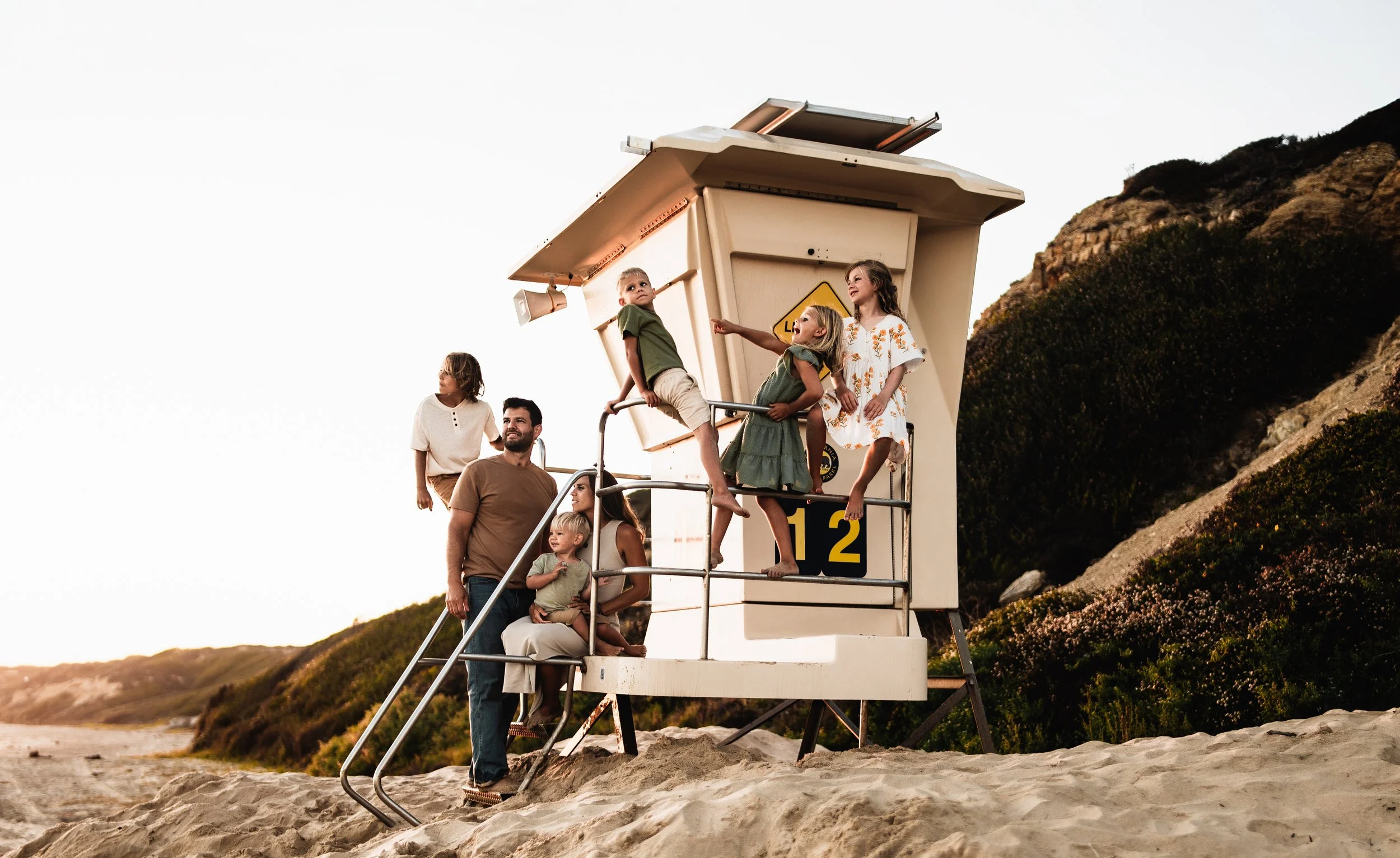 A lifeguard tower on a sandy beach with children and a man sitting and standing on it, with a hill covered in grass and shrubs in the background.