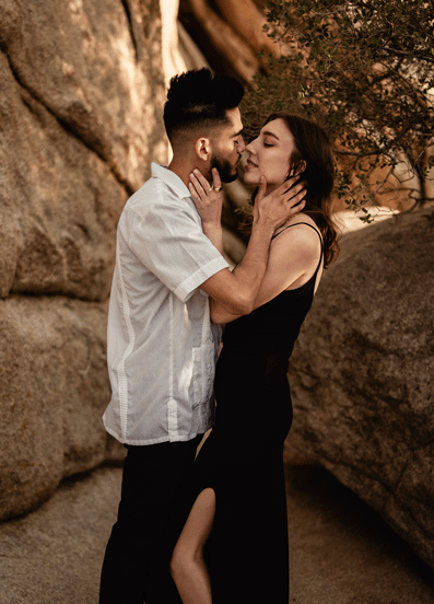 Couple kissing in a rocky outdoor setting, woman in a black dress, and man in a white shirt.