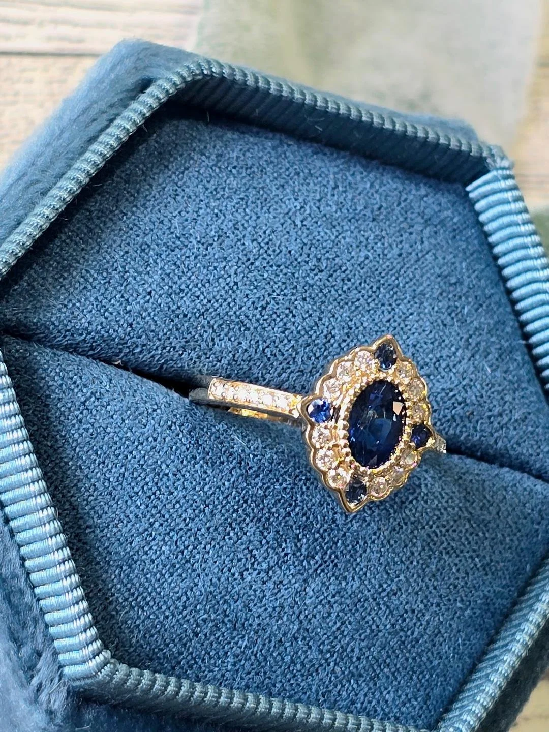Sideview view of a sapphire ring with vintage styled halo in a blue box on white wooden background.