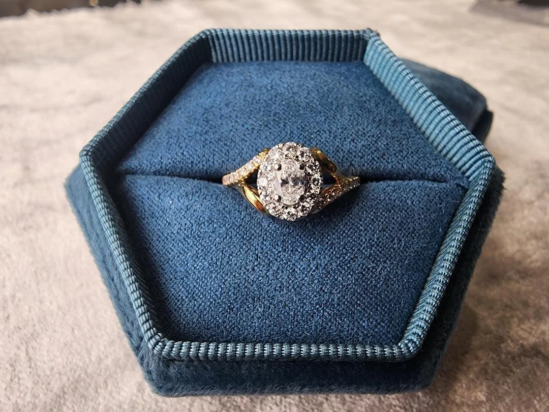 yellow gold diamond ring with a oval diamond with a halo and split shoulders in a blue ring box with  grey fabric in background
