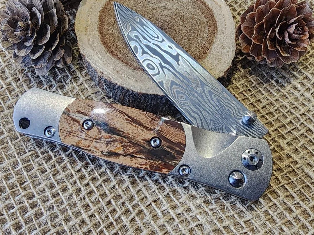 open damascus folding pocket knife with button lock and mammoth tusk ivory on burlap with wood slice and pinecone in background