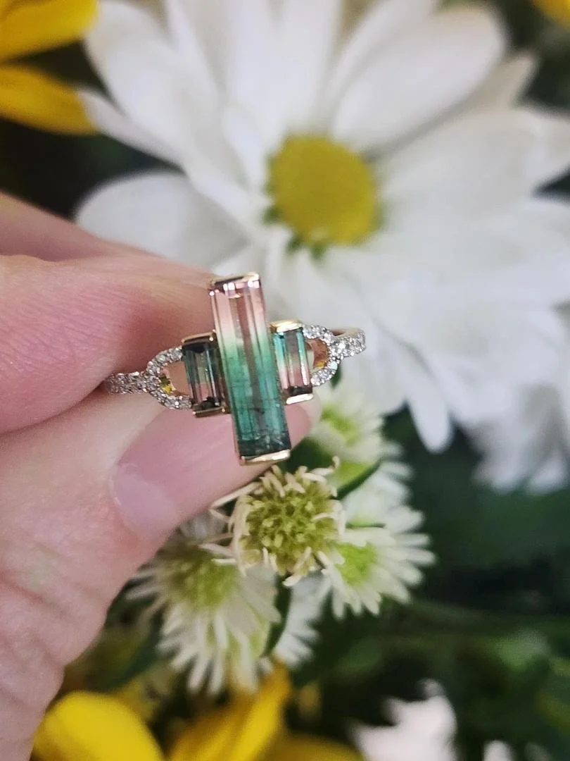 finger holding a Yellow gold ring with 3 stone watermelon tourmaline and diamonds with spring flowers in background