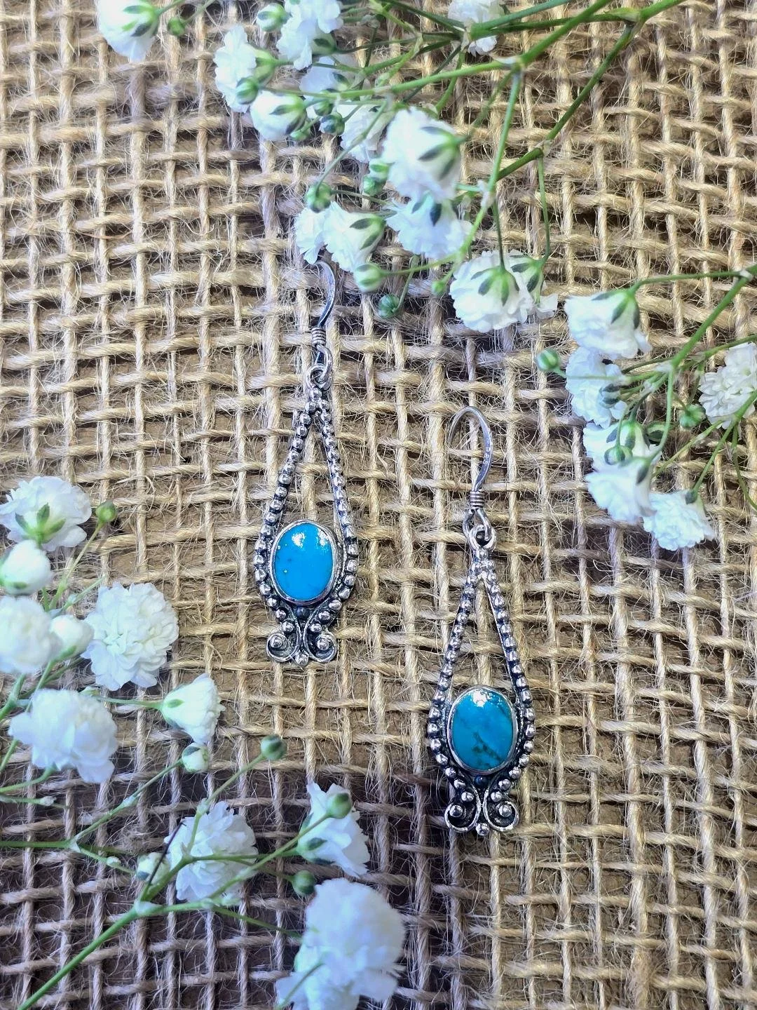 Front view of silver turquoise dangle earrings on a burlap background with white flowers on the corners