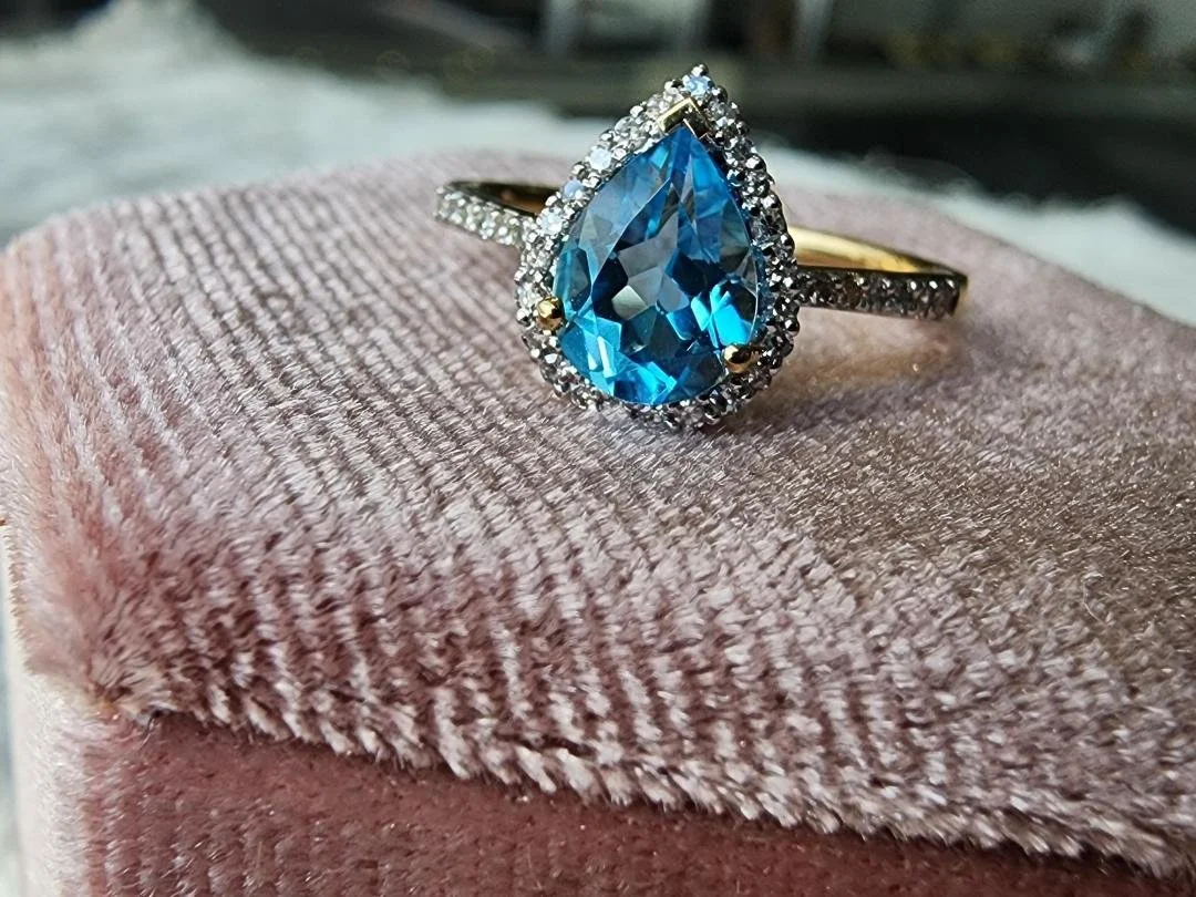yellow gold ring with pear shaped blue topaz and diamond halo and shoulders accents on a pink box