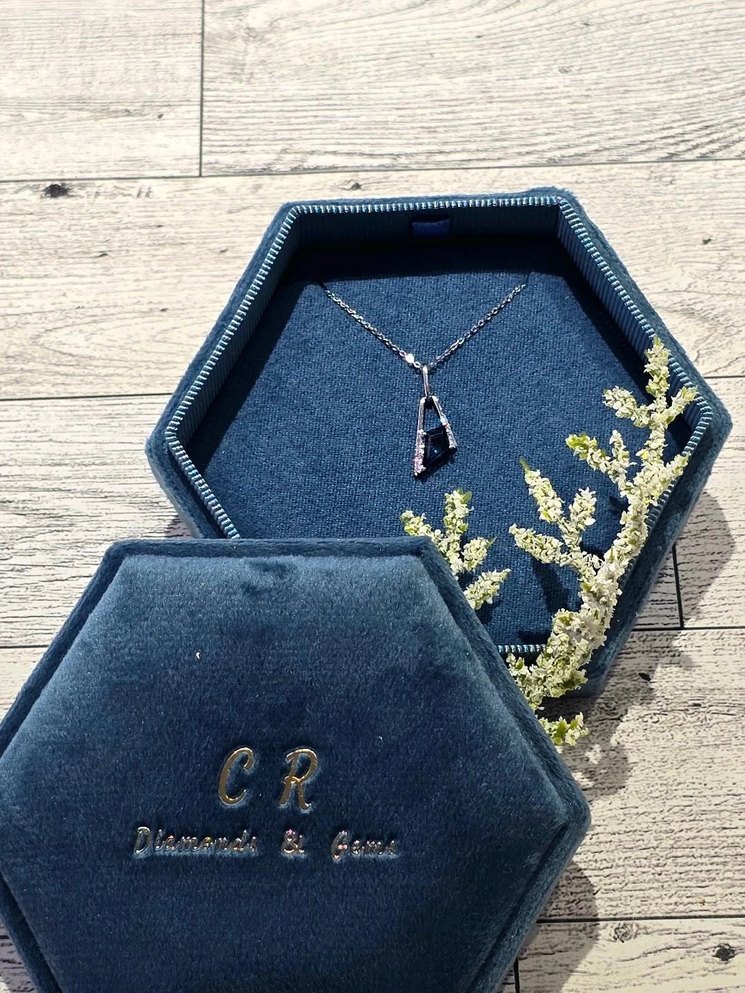 Front view of white gold London blue topaz necklace in a navy blue box on a white wooden background