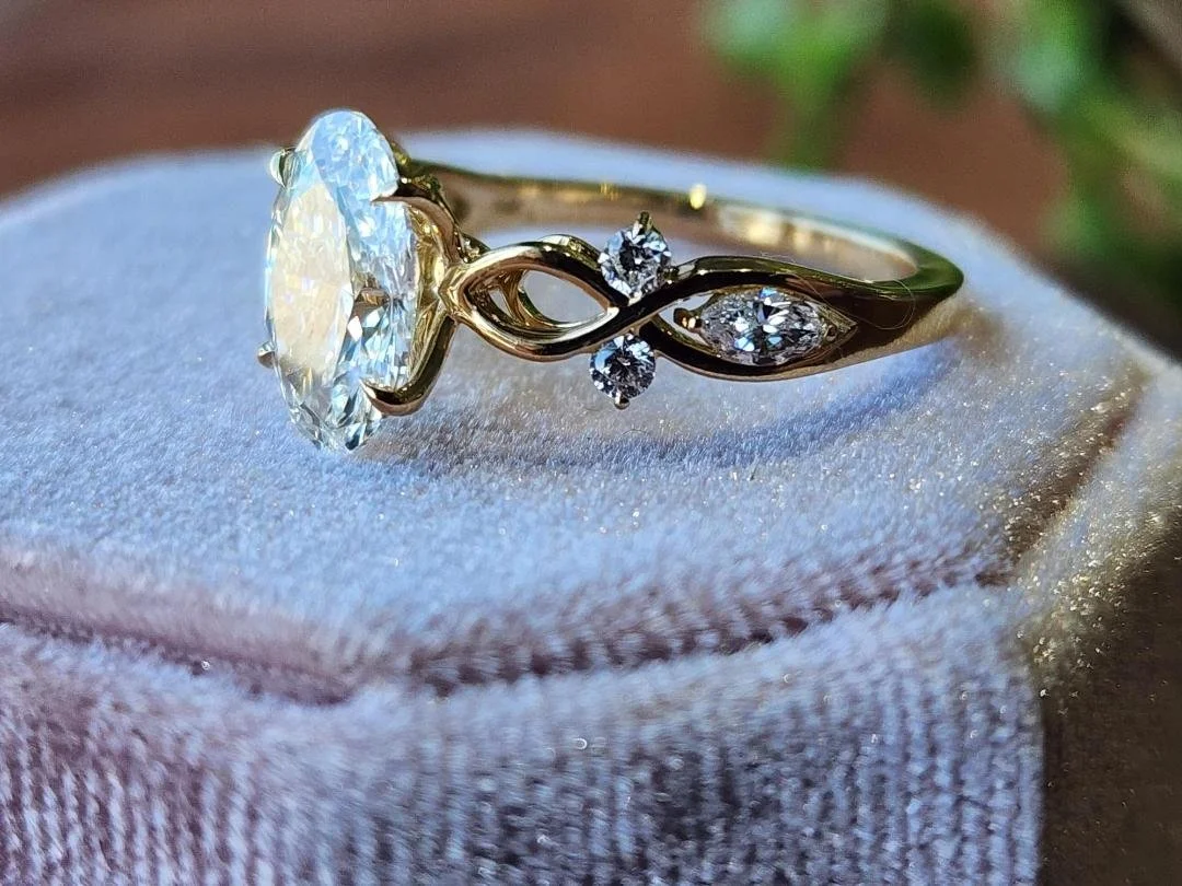 Oval lab grown diamond with rounds and marquise on the sides in yellow gold with a pink box and brown and green in background
