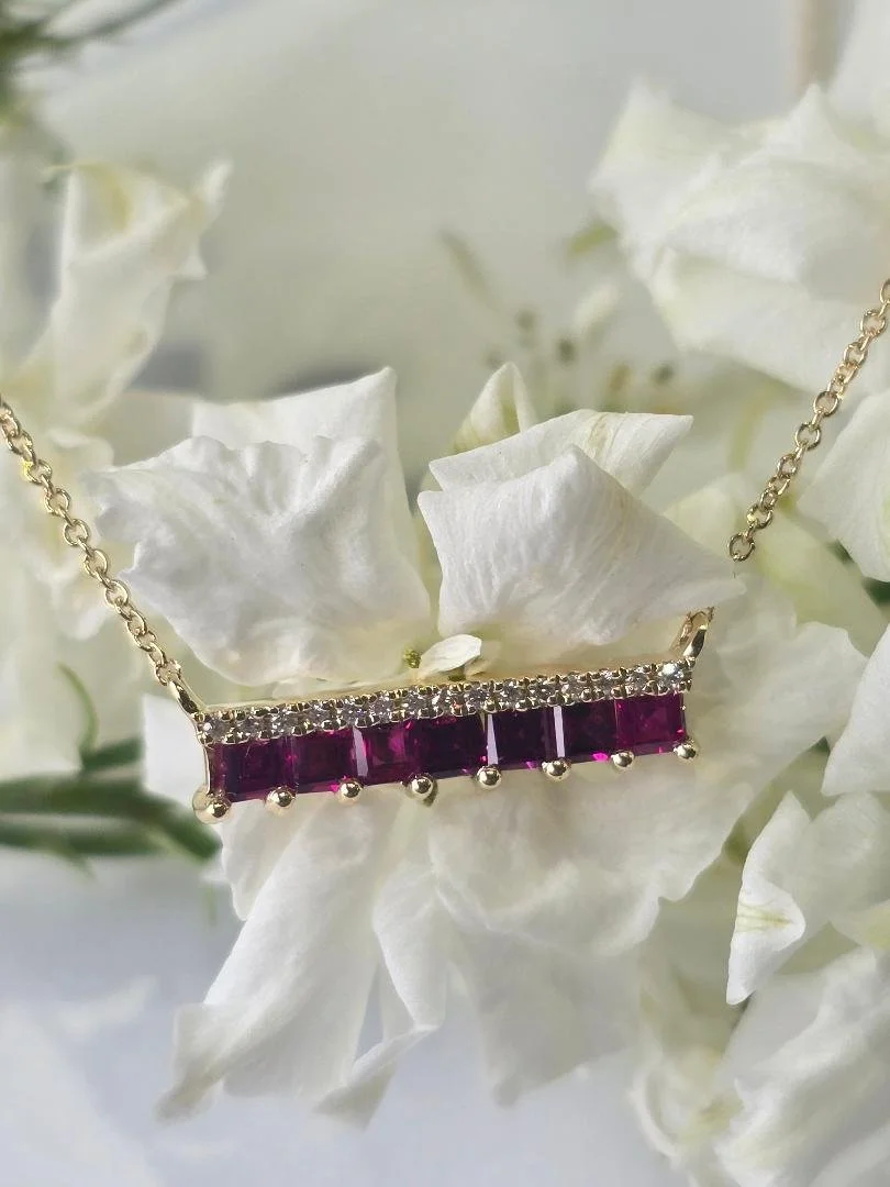 Front view of a yellow gold ruby and diamond bar style pendant draped over white flowers