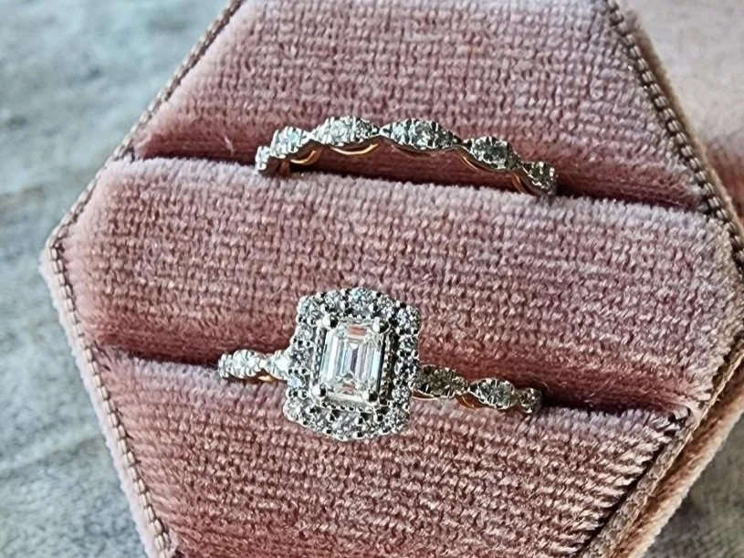 White and rose gold diamond wedding ring set with a emerald cut center diamond in a halo of round diamonds in a pink box on a grey background