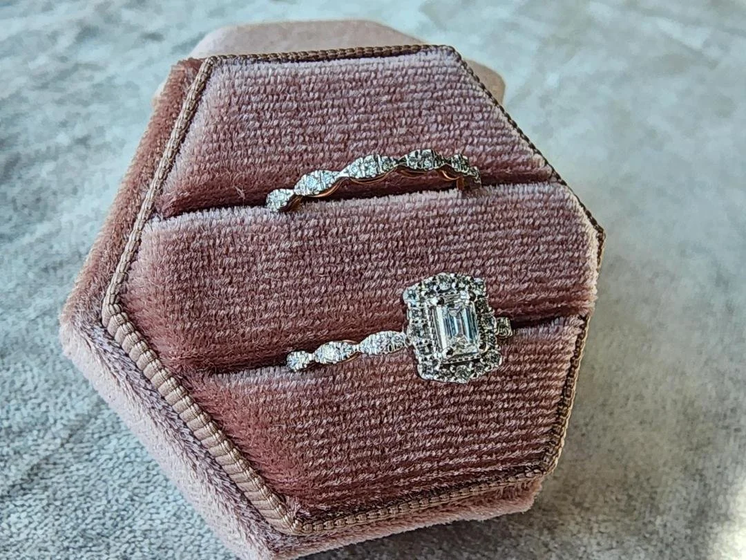 White and rose gold diamond wedding ring set with a emerald cut center diamond in a halo of round diamonds in a pink box on a grey background