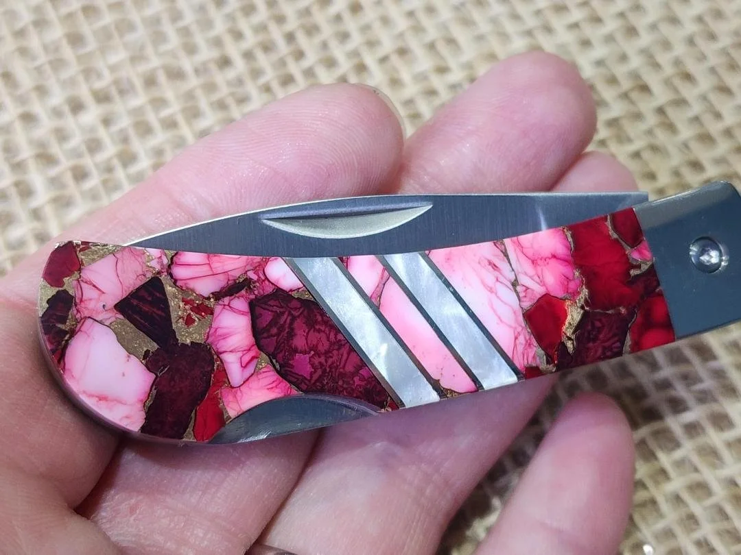 hand holding 3" knife the "red dahlia" with pink to red inlay and copper veining.  Then two mother of pearl stripes in the center on wood display with burlap in background
