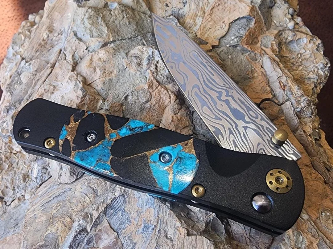 open black folding knife with damascus blade with turquoise onyx and copper on the handle on rock