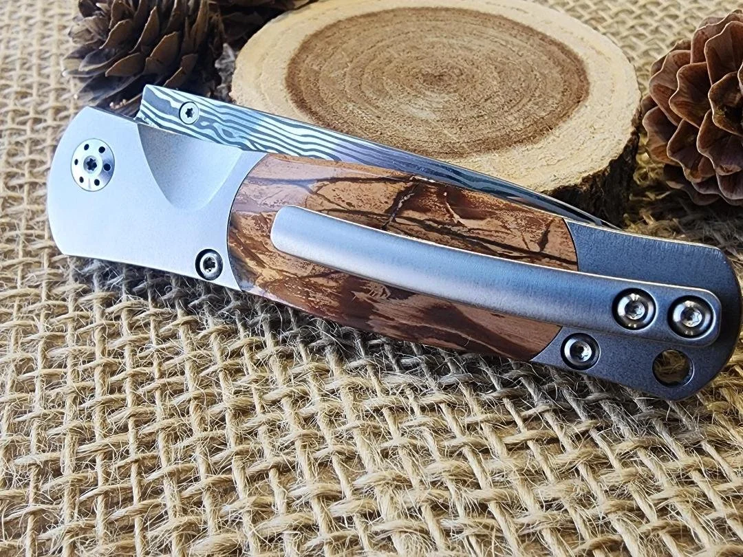 damascus folding pocket knife with button lock and mammoth tusk ivory  with clip on burlap with wood slice and pinecone in background