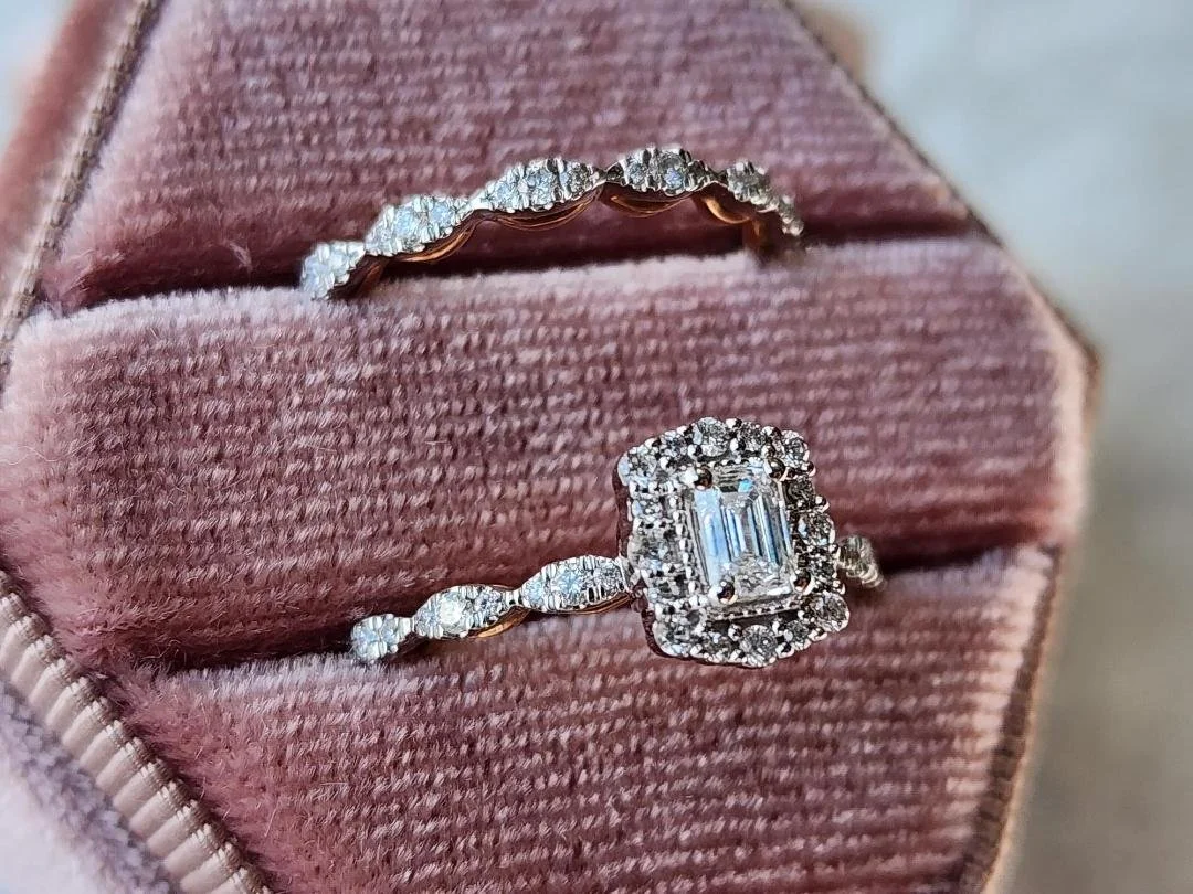 side view of White and rose gold diamond wedding ring set with a emerald cut center diamond in a halo of round diamonds in a pink box on a grey background