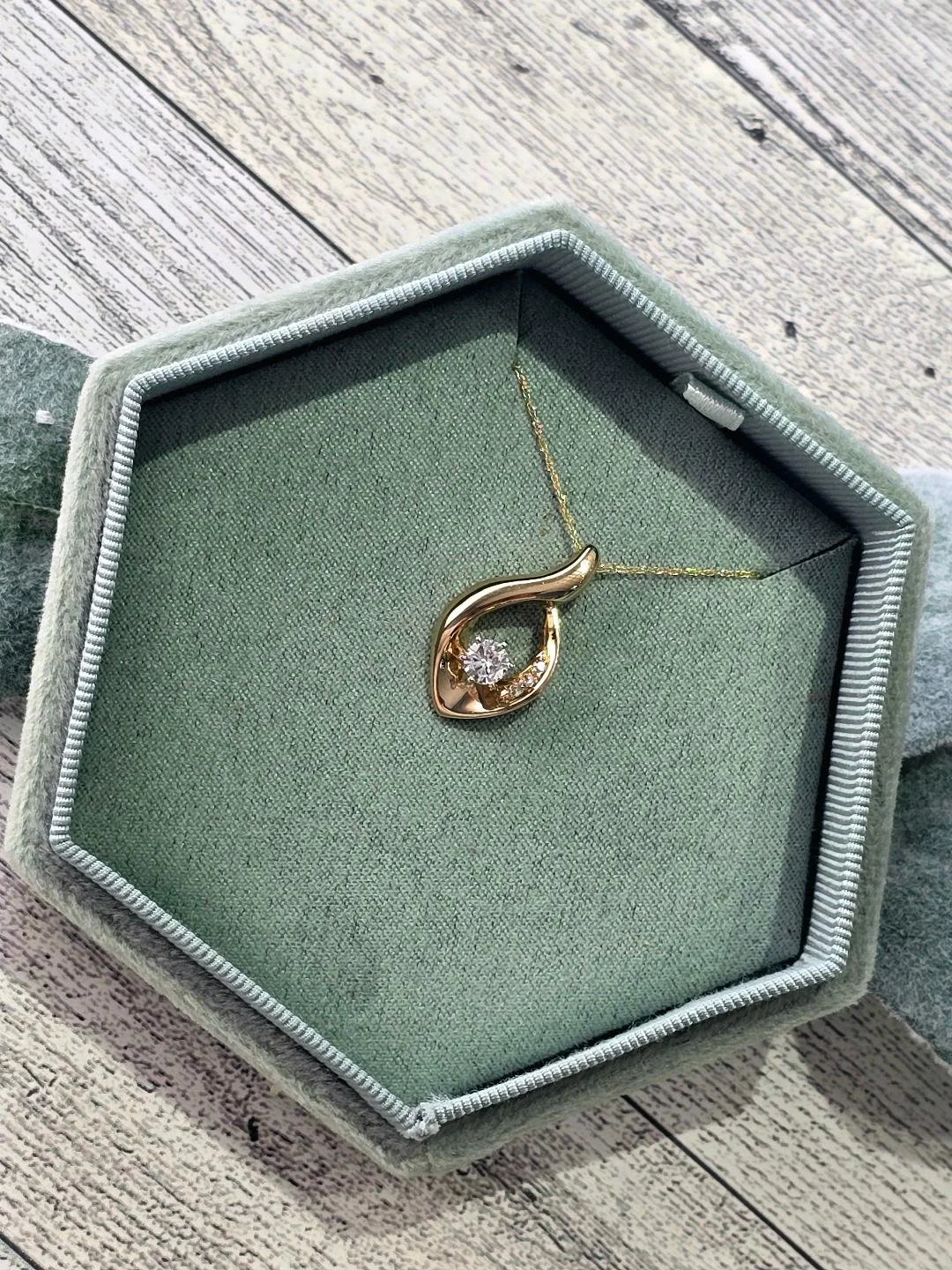 Angled view of yellow gold drop pendant in a sage green box with greenery around the box