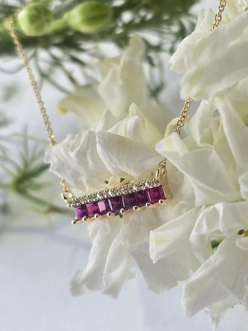 angled view of a yellow gold ruby and diamond bar style pendant draped over white flowers