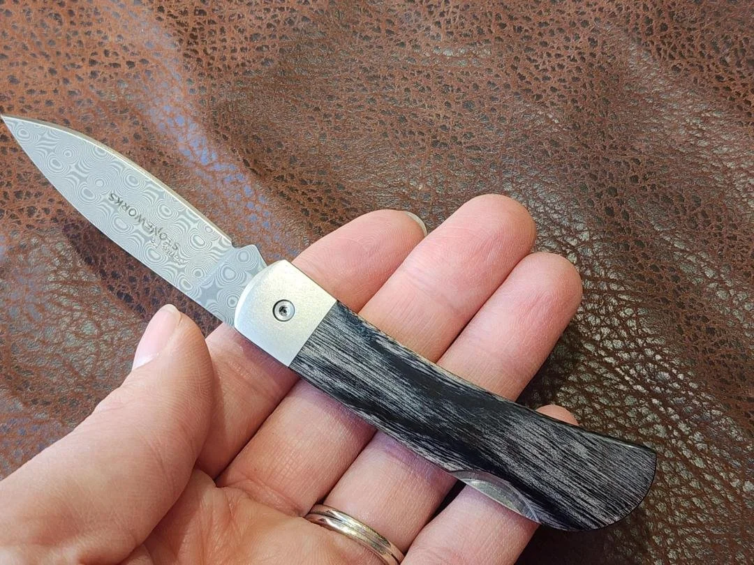 hand holding 3" knife with raindrop damascus blade and black onyx and agatized dinosaur bone inlay on a leather style background