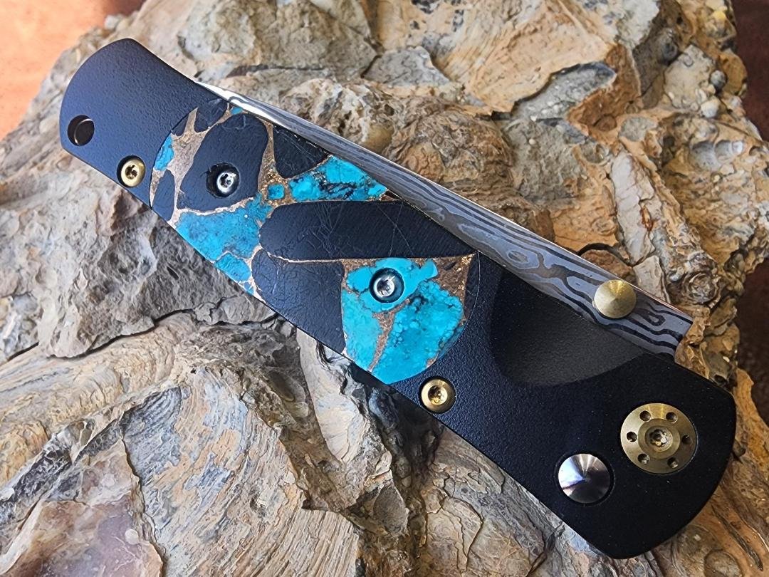 black folding knife with damascus blade with turquoise onyx and copper on the handle on rock