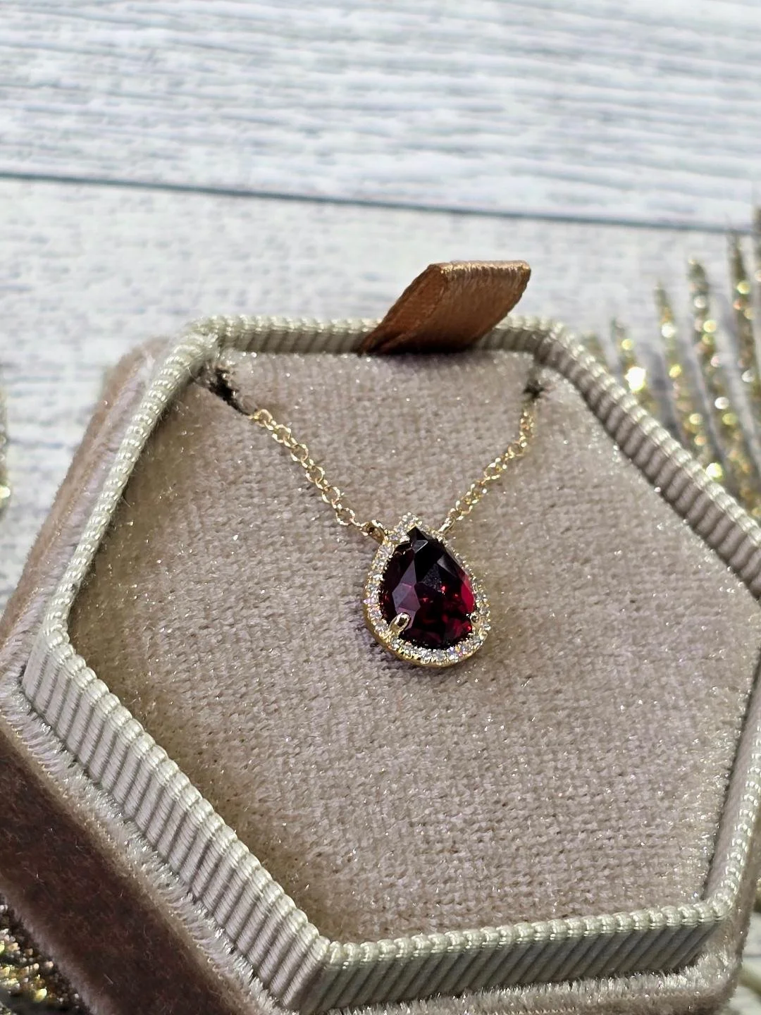 Angled view of a yellow gold garnet and diamond stationary pendant in a brown box with a white wood background