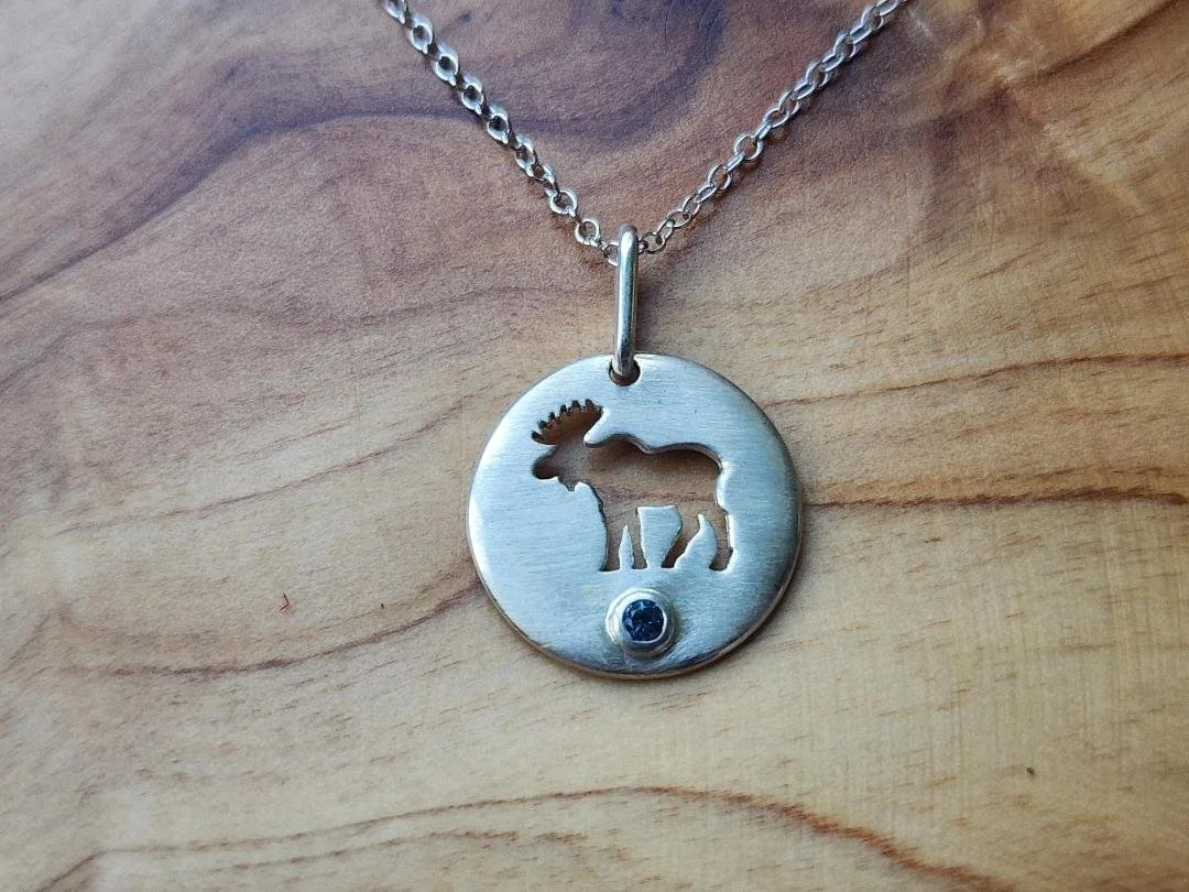Round silver necklace with moose cut out and round blue sapphire with a wood background