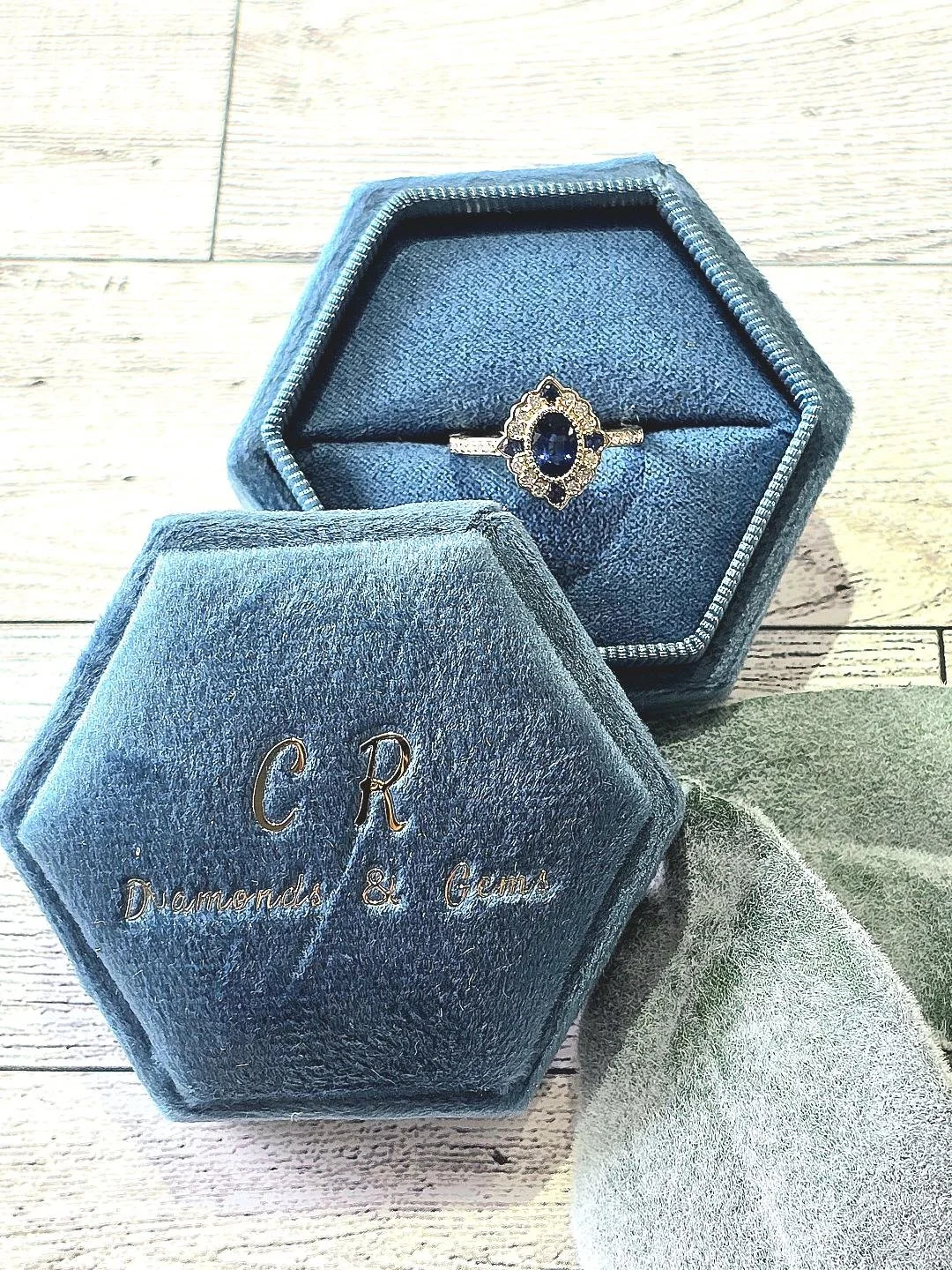 Front view of a shapphire ring with vintage styled halo in a blue box on white wooden background.