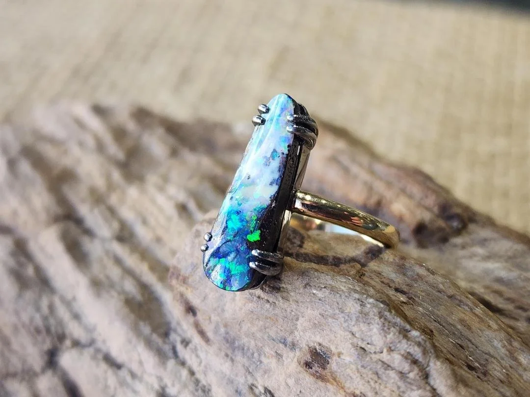 side view of Blue and white elongated Australian boulder opal ring in blackened sterling silver and yellow gold on a rock with burlap background