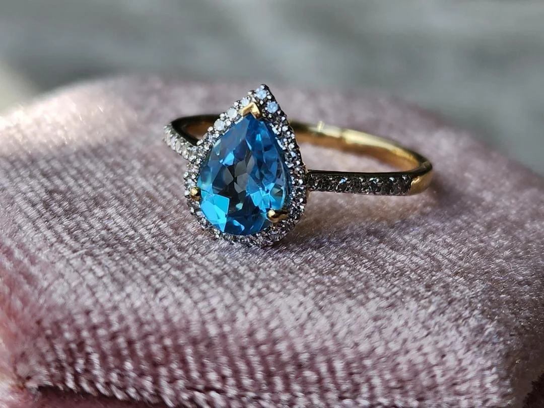 yellow gold ring with pear shaped blue topaz and diamond halo and shoulders accents on a pink box