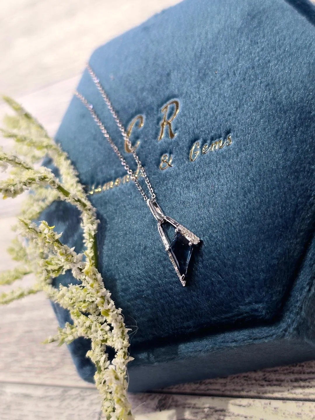 Angled view of white gold London blue topaz necklace in a navy blue box on a white wooden background