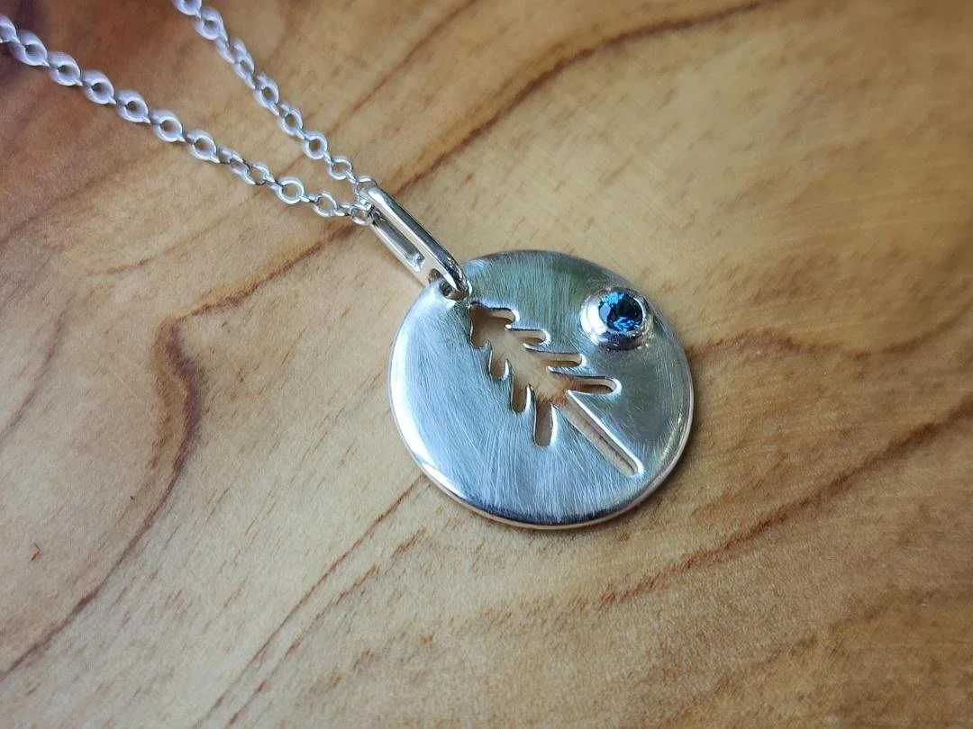 side view of sterling silver round disk pendant with a tree cutout and a round blue sapphire on wood