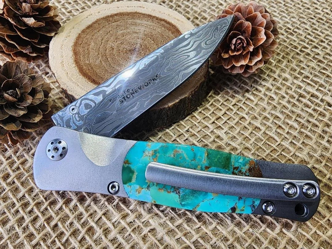 open One 3.5" damascus knife with a turquoise inlay with a clip on end of knife. on burlap with a wood slice and pinecones in background