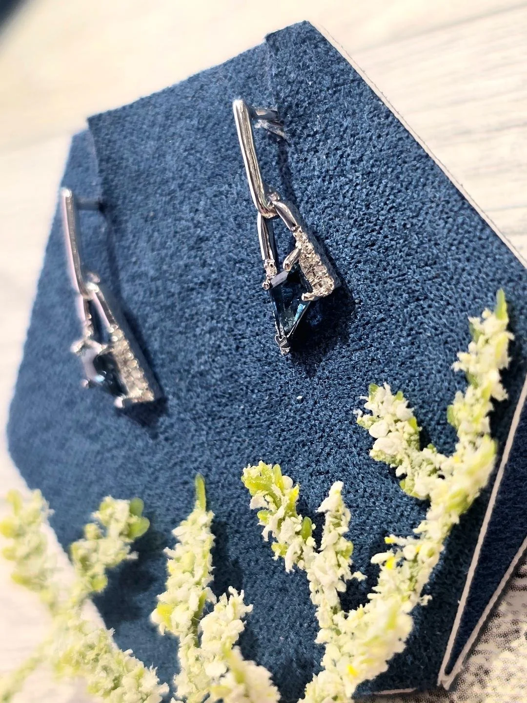 Side view of white gold London blue topaz earrings in a navy blue box with a white wooden background