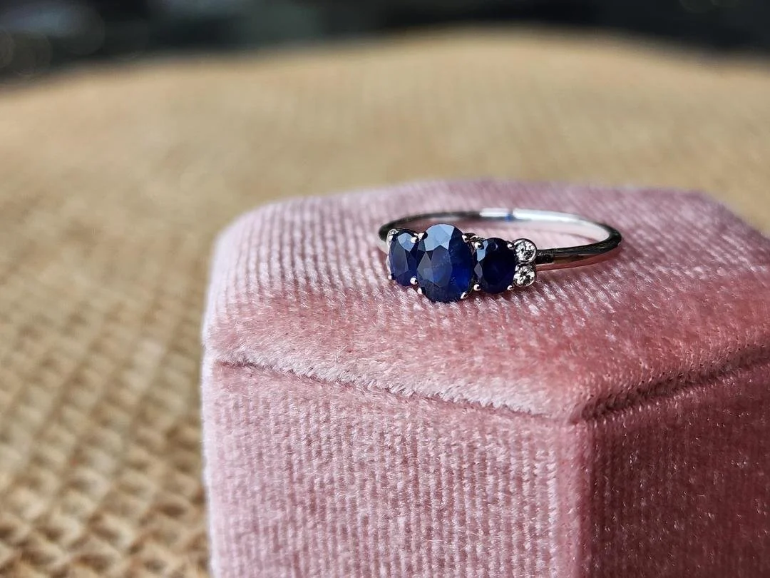 white gold 3 stone sapphire and diamond accent ring on a pink box with a brown background