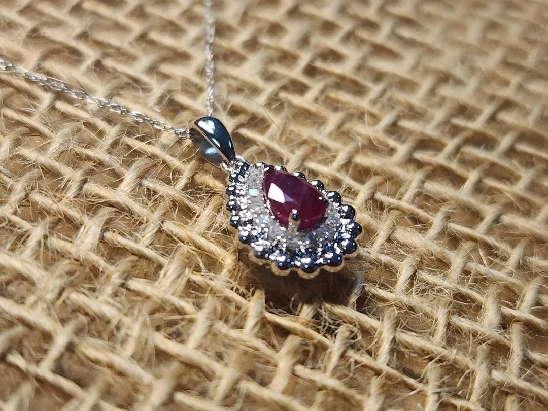 side view White gold necklace with a pear shaped red ruby with a halo of round diamonds and a white gold bead texture on a burlap background