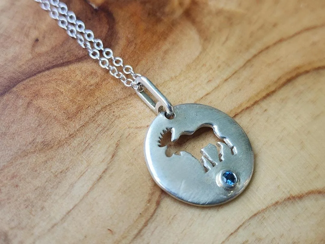 side view Round silver necklace with moose cut out and round blue sapphire with a wood background