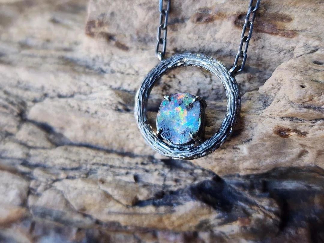 Blackened sterling silver necklace with a circle pendant featuring a round boulder opal displayed on a rock