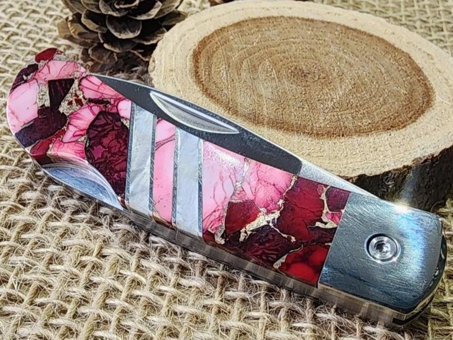 3" knife the "red dahlia" with pink to red inlay and copper veining.  Then two mother of pearl stripes in the center on wood display with pinecones and burlap in background