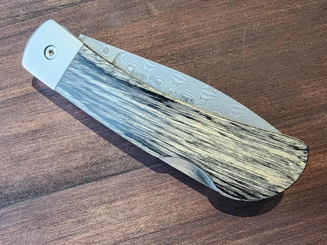 Closed Damascus folding pocketknife with stabilized eastern birch handle and raindrop Damascus blade with wooden background